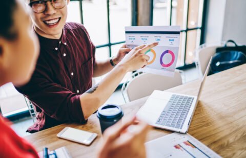 From above of smart smiling young coworkers in casual apparel talking with each other surfing on laptop analyzing graph in creative workplace at table with cup of coffee